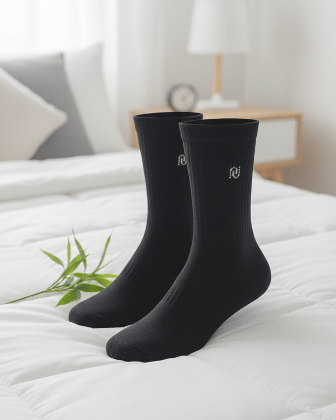 Two black bamboo men’s crew socks standing upright on a white bed, featuring a stitched minimalist logo, styled in a bright modern bedroom.