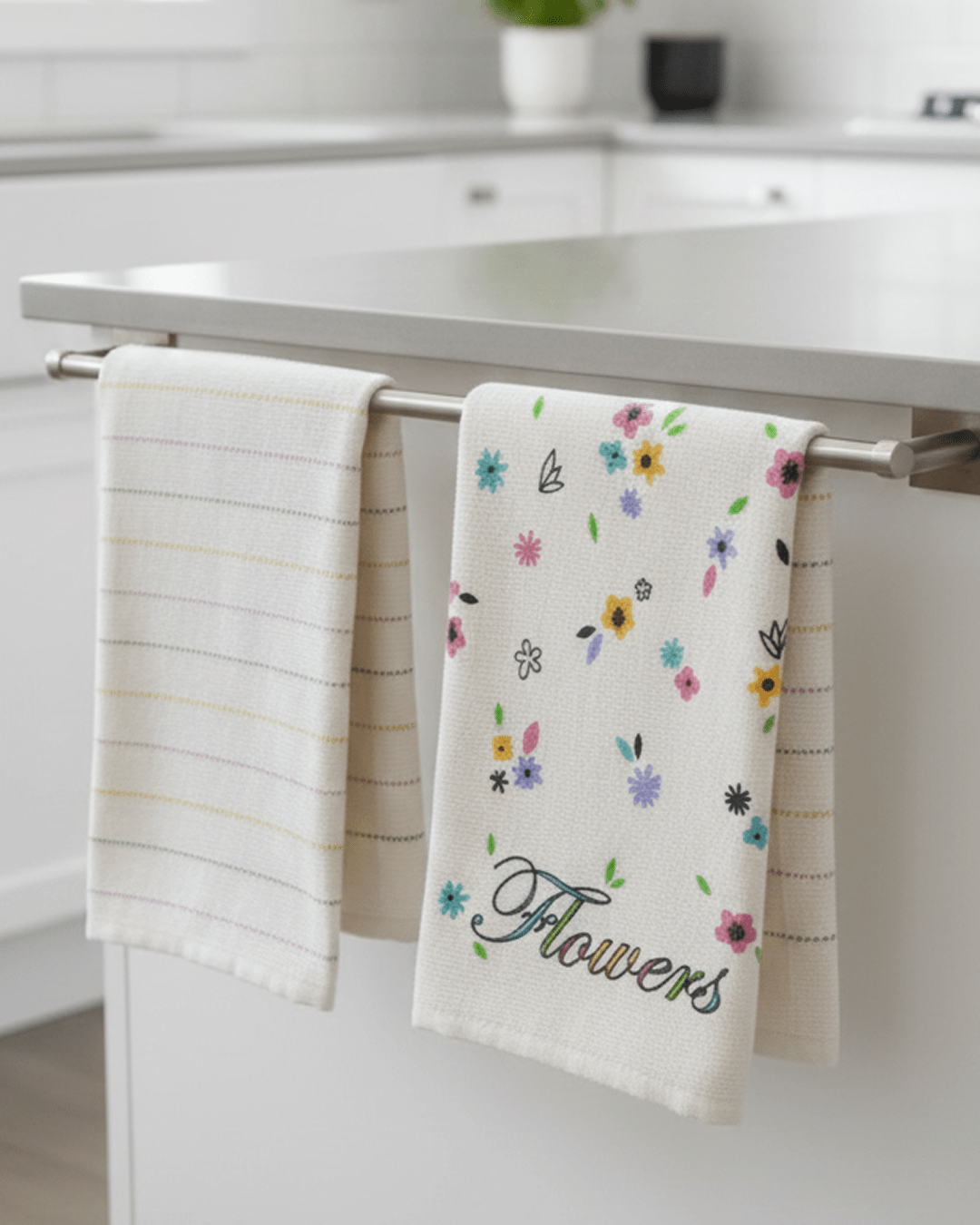 Two kitchen towels hanging on a stainless steel bar in a modern white kitchen, one with colorful floral patterns and the word “Flowers,” the other with subtle pastel stripes.