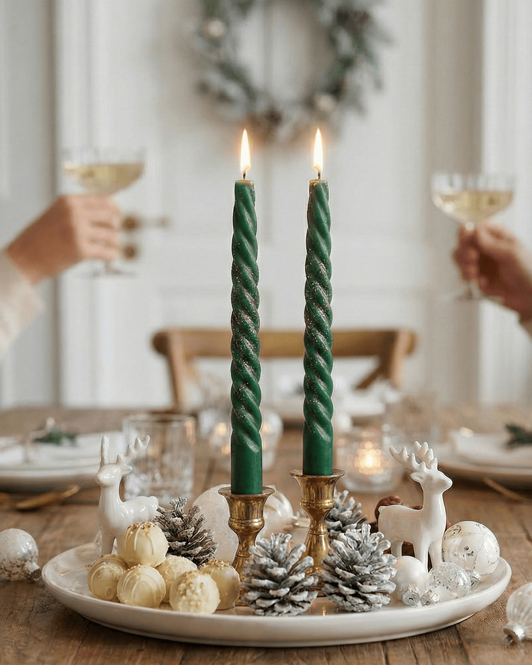 A pair of long, silver twisted taper candles burning in brass holders, forming the centerpiece of a festive holiday table setting with white reindeer figurines and snow-dusted pinecones.