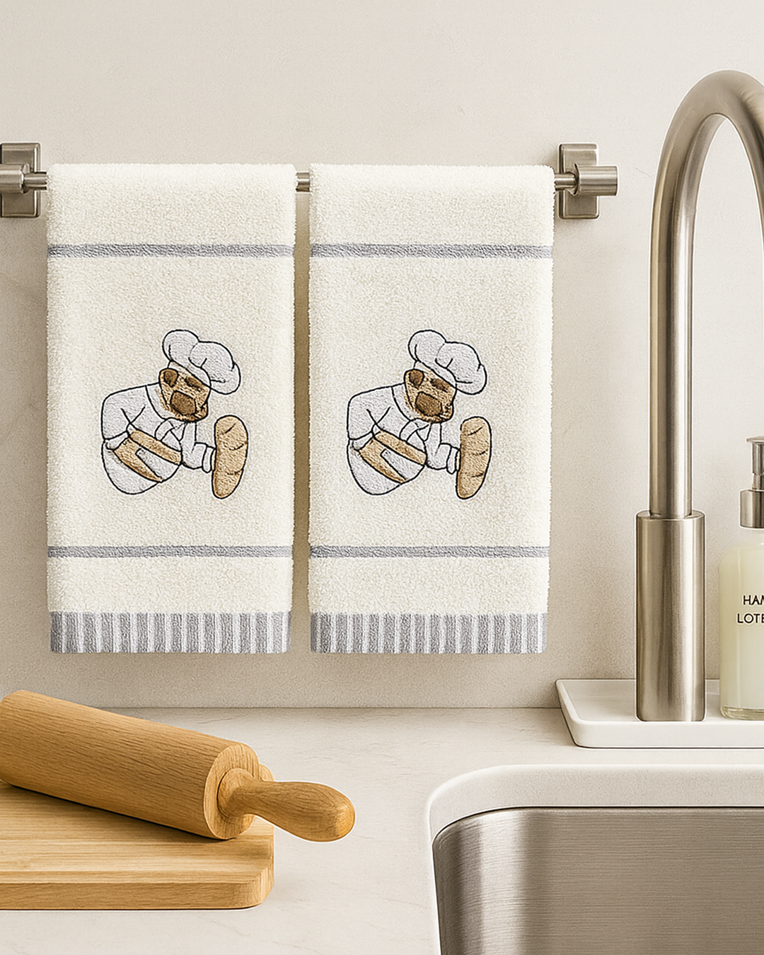 Two white cotton kitchen towels with embroidered chef holding bread, hanging on a metal towel bar above a kitchen sink with wooden rolling pin and soap dispenser.