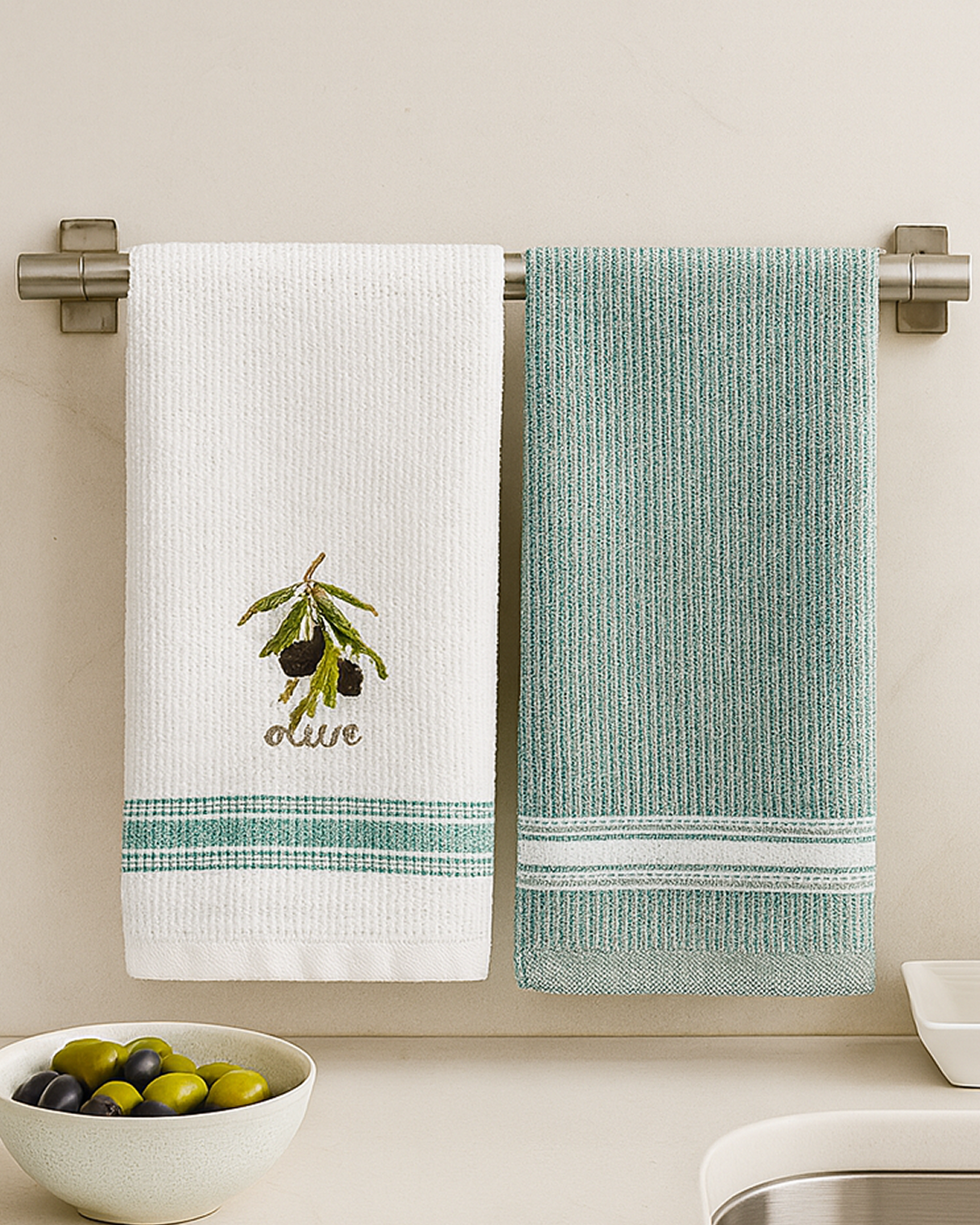 Two white cotton kitchen towels with embroidered olive, hanging on a metal towel bar above a kitchen sink with wooden rolling pin and soap dispenser.