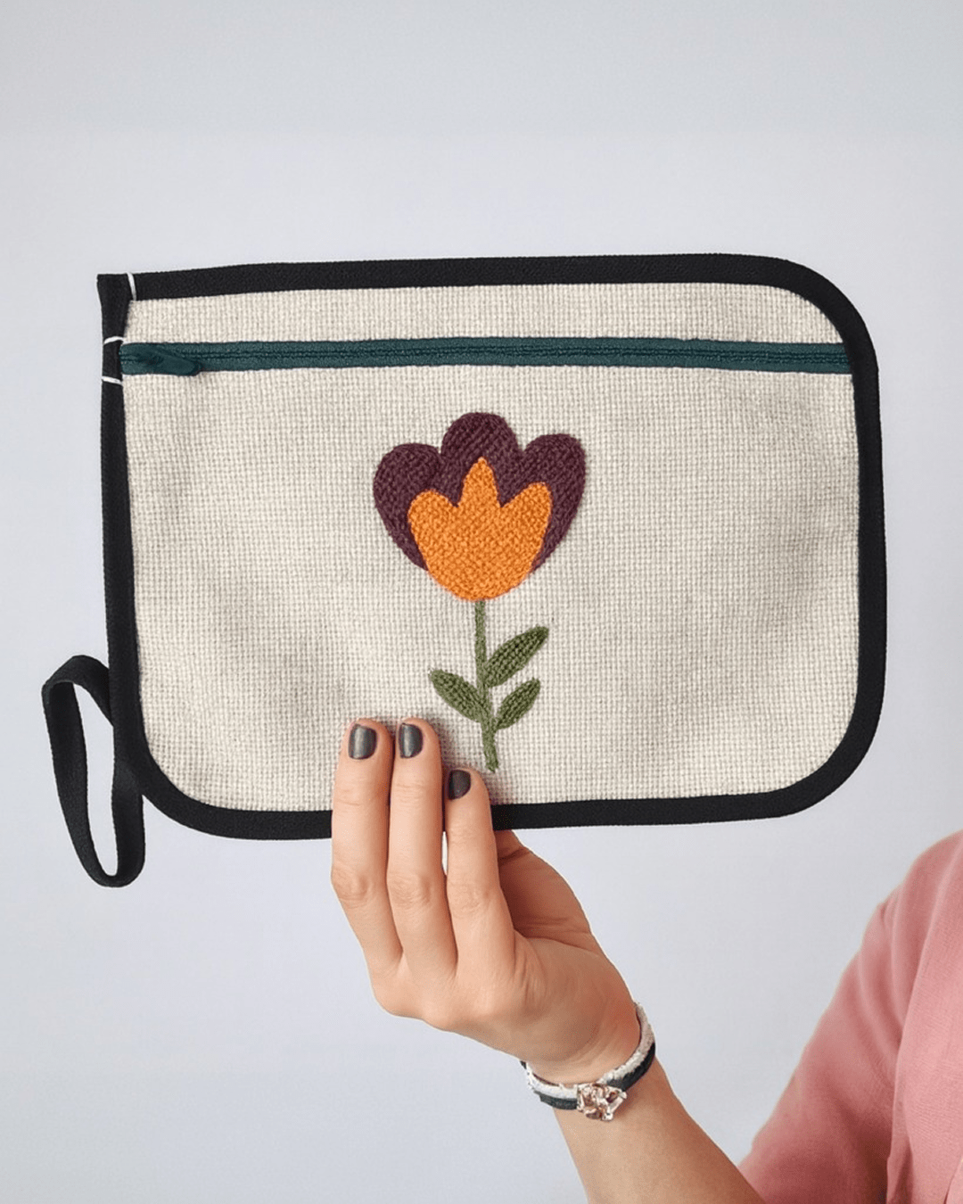 Punch needle embroidered pouch with floral design held by hand, cream textured fabric with black border and zipper.