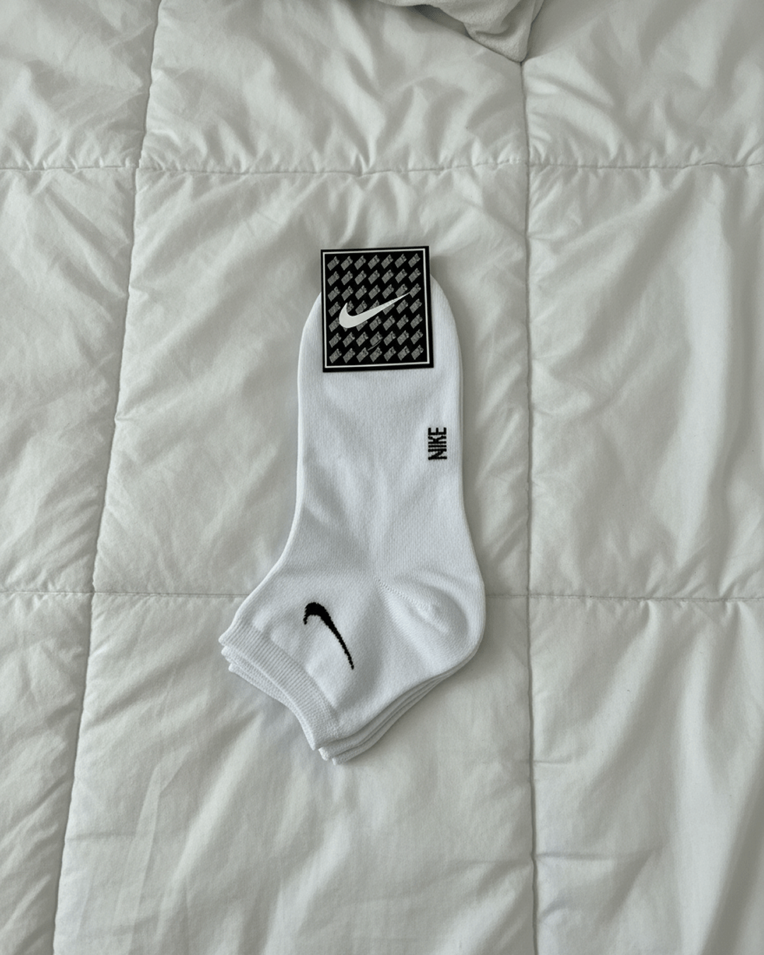 A white multi-pack of Nike ankle socks made from 100% Turkish cotton, featuring the black Nike swoosh and logo, resting on a white quilted bedspread.