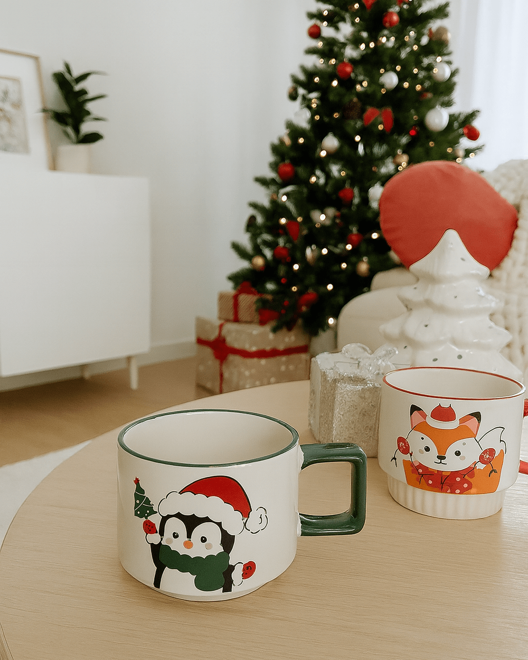 A set of two ceramic Christmas mugs on a wooden table: one features a penguin in a Santa hat, and the other features a fox in a festive scarf, with a decorated Christmas tree in the background.